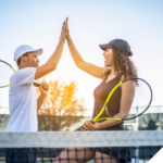 Ein Mann und eine Frau geben sich auf einem Tennisplatz ein High-Five, während sie Tennisschläger in der Hand halten. Beide tragen sportliche Kleidung und Kappen, und die Szene wird von warmem Sonnenlicht im Hintergrund erhellt. Das High-Five symbolisiert die Energie, Vitalität und den Teamgeist, die durch eine bewusste Lebensweise und Ernährung – wie der veganen Ernährung – gefördert werden können. Es unterstreicht die Aussage des Titels **"Sportliche Vorteile durch vegane Ernährung – Wissenschaftliche Einblicke und Erfahrungsberichte"**, indem es aufzeigt, wie eine ausgewogene pflanzliche Ernährung positive Auswirkungen auf Leistungsfähigkeit und Wohlbefinden im Sport haben kann. In der unteren rechten Ecke befindet sich das Logo von **"Vegane Sporternährung für alle" by Anne Mayer**, dargestellt durch ein grünes Brokkoli-Icon.