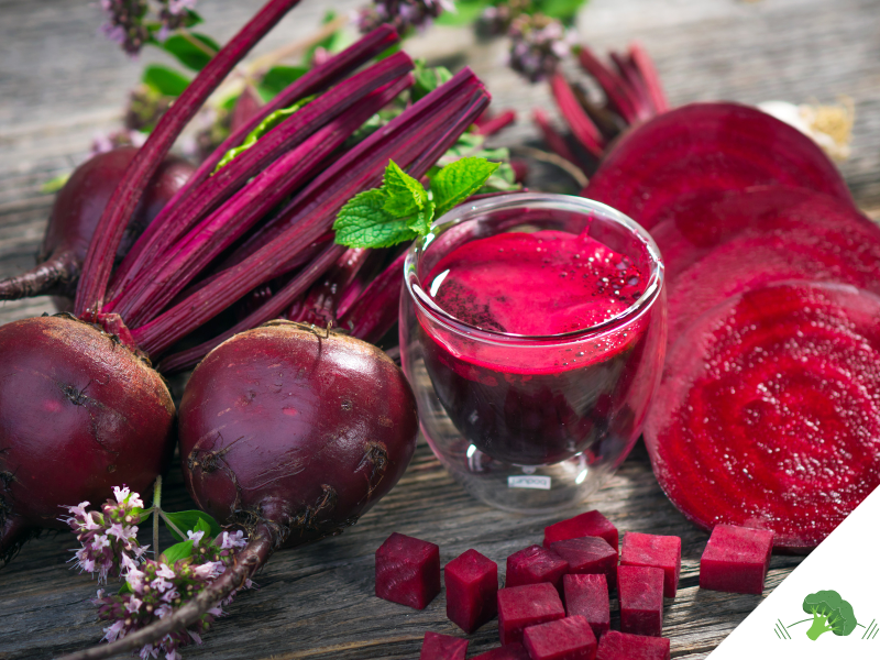 Frische rote Bete mit Blättern und Stängeln, daneben geschnittene Scheiben und Würfel sowie ein Glas frisch gepresster Rote-Bete-Saft, garniert mit einem Minzblatt, auf einer rustikalen Holzoberfläche arrangiert. In der unteren rechten Ecke befindet sich das Logo von "Vegane Sporternährung für alle" by Anne Mayer, dargestellt durch ein grünes Brokkoli-Icon.