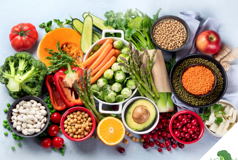 Eine bunte Auswahl an frischen, pflanzlichen Lebensmitteln, darunter Brokkoli, Kürbis, Karotten, Rosenkohl, Avocado, Tomaten, Paprika, Bohnen, Linsen, Cranberries und Orangen. Alles ist dekorativ auf einem hellen Untergrund arrangiert, um eine gesunde und nährstoffreiche Ernährung zu symbolisieren.