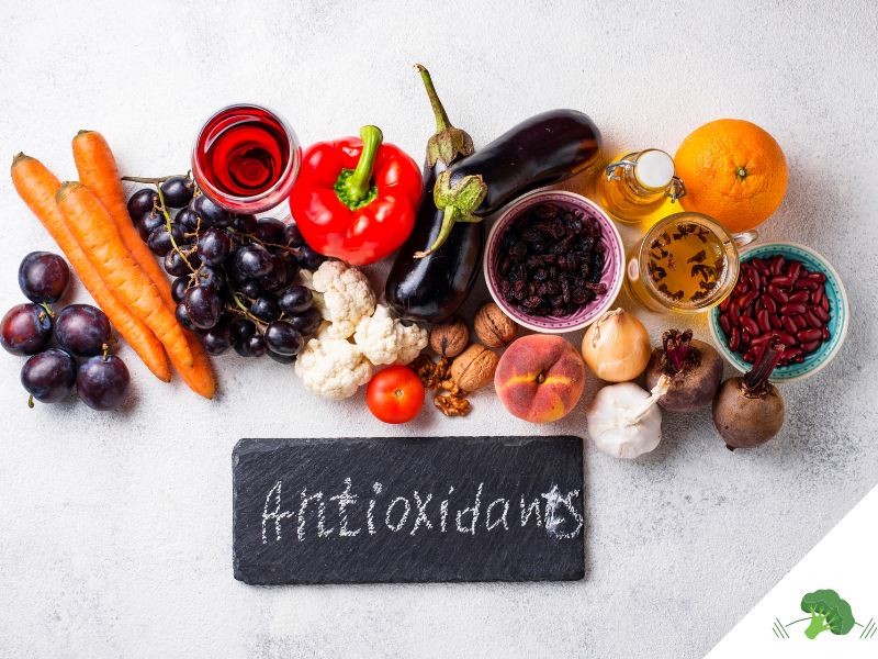 Eine Auswahl antioxidativer Lebensmittel: Eine Tafel mit der Aufschrift 'Antioxidants' liegt umgeben von verschiedenen farbenfrohen Nahrungsmitteln, darunter Karotten, Trauben, Paprika, Blumenkohl, Aubergine, getrocknete Früchte, Walnüsse, Tomaten, Orangen, rote Bohnen und Rote Bete. Im Hintergrund sind auch ein Glas roter Saft und eine Flasche Öl zu sehen. Diese Darstellung symbolisiert antioxidative Ernährung