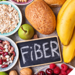 Eine Auswahl ballaststoffreicher Lebensmittel: Eine Tafel mit der Aufschrift 'Fiber' liegt umgeben von verschiedenen Nahrungsmitteln, darunter Bananen, Brot, Haferflocken, eine grüne Birne, rote und weiße Bohnen, Walnüsse, Himbeeren, Kirschen und Brokkoli. Diese symbolisieren eine gesunde und ballaststoffreiche Ernährung.