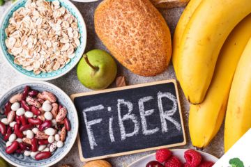 Eine Auswahl ballaststoffreicher Lebensmittel: Eine Tafel mit der Aufschrift 'Fiber' liegt umgeben von verschiedenen Nahrungsmitteln, darunter Bananen, Brot, Haferflocken, eine grüne Birne, rote und weiße Bohnen, Walnüsse, Himbeeren, Kirschen und Brokkoli. Diese symbolisieren eine gesunde und ballaststoffreiche Ernährung.