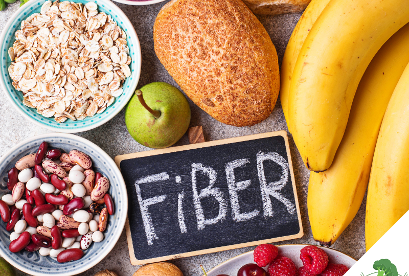 Eine Auswahl ballaststoffreicher Lebensmittel: Eine Tafel mit der Aufschrift 'Fiber' liegt umgeben von verschiedenen Nahrungsmitteln, darunter Bananen, Brot, Haferflocken, eine grüne Birne, rote und weiße Bohnen, Walnüsse, Himbeeren, Kirschen und Brokkoli. Diese symbolisieren eine gesunde und ballaststoffreiche Ernährung.