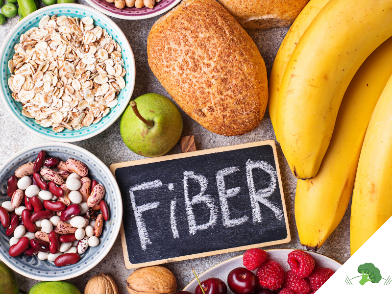 Eine Auswahl ballaststoffreicher Lebensmittel: Eine Tafel mit der Aufschrift 'Fiber' liegt umgeben von verschiedenen Nahrungsmitteln, darunter Bananen, Brot, Haferflocken, eine grüne Birne, rote und weiße Bohnen, Walnüsse, Himbeeren, Kirschen und Brokkoli. Diese symbolisieren eine gesunde und ballaststoffreiche Ernährung.