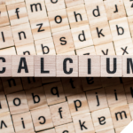 Holzklötze mit schwarzen Buchstaben darauf formen das Wort „CALCIUM“. Die Klötze liegen auf einer Fläche mit weiteren Holzklötzen, die verschiedene Buchstaben tragen.