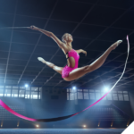 Eine Athletin in einem pinken Outfit führt einen dynamischen Sprung in einer großen Sporthalle aus, mit einem rhythmischen Band in der Hand, das in einer geschwungenen Bewegung hinter ihr herläuft. Das Bild symbolisiert optimierte Performance, Präzision und sportliche Höchstleistungen.