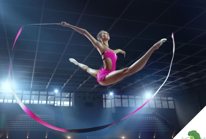 Eine Athletin in einem pinken Outfit führt einen dynamischen Sprung in einer großen Sporthalle aus, mit einem rhythmischen Band in der Hand, das in einer geschwungenen Bewegung hinter ihr herläuft. Das Bild symbolisiert optimierte Performance, Präzision und sportliche Höchstleistungen.