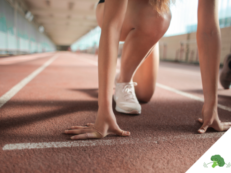 Nahaufnahme einer startenden Läuferin auf einer Laufbahn, die Hände an der Startlinie. Im Hintergrund ist eine helle, moderne Indoor-Sporthalle zu sehen.