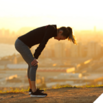 Frau in Sportkleidung steht erschöpft mit den Händen auf den Knien auf einem Hügel, im Hintergrund eine Stadtlandschaft im Sonnenaufgang.