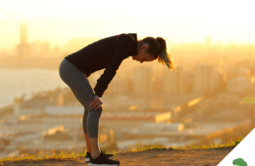 Frau in Sportkleidung steht erschöpft mit den Händen auf den Knien auf einem Hügel, im Hintergrund eine Stadtlandschaft im Sonnenaufgang.