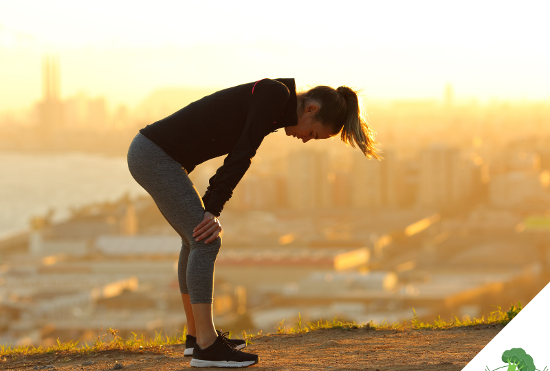 Frau in Sportkleidung steht erschöpft mit den Händen auf den Knien auf einem Hügel, im Hintergrund eine Stadtlandschaft im Sonnenaufgang.