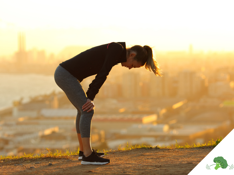 Frau in Sportkleidung steht erschöpft mit den Händen auf den Knien auf einem Hügel, im Hintergrund eine Stadtlandschaft im Sonnenaufgang.