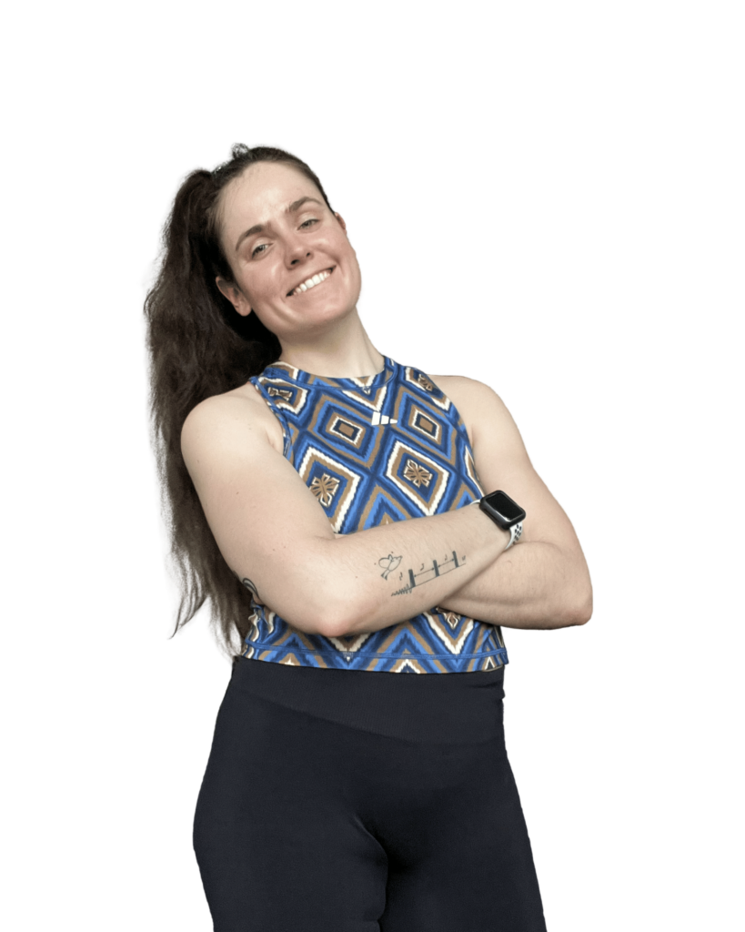 Eine Frau mit langem, dunkelbraunem Haar trägt ein ärmelloses Oberteil mit blau-goldenem geometrischem Muster und schwarze Sportleggings. Sie steht mit verschränkten Armen, lächelt selbstbewusst in die Kamera und trägt eine Smartwatch am linken Handgelenk. Auf ihrem rechten Unterarm ist ein kleines Tattoo zu sehen. Der Hintergrund ist entfernt.