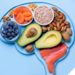 Gesunde Lebensmittel in der Form eines Gehirns angeordnet, darunter Avocado, Blaubeeren, Trockenaprikosen, Nüsse, Sonnenblumenkerne und Lachs, symbolisieren den Zusammenhang zwischen Ernährung und Gehirnfunktion.