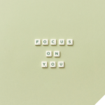 Scrabble-Buchstaben auf einem hellgrünen Hintergrund formen die Worte „FOCUS ON YOU“ in drei Zeilen, unten rechts befindet sich ein freier Bereich mit weißem Dreieck und einer Illustration eines grünen Brokkoli mit Hanteln.