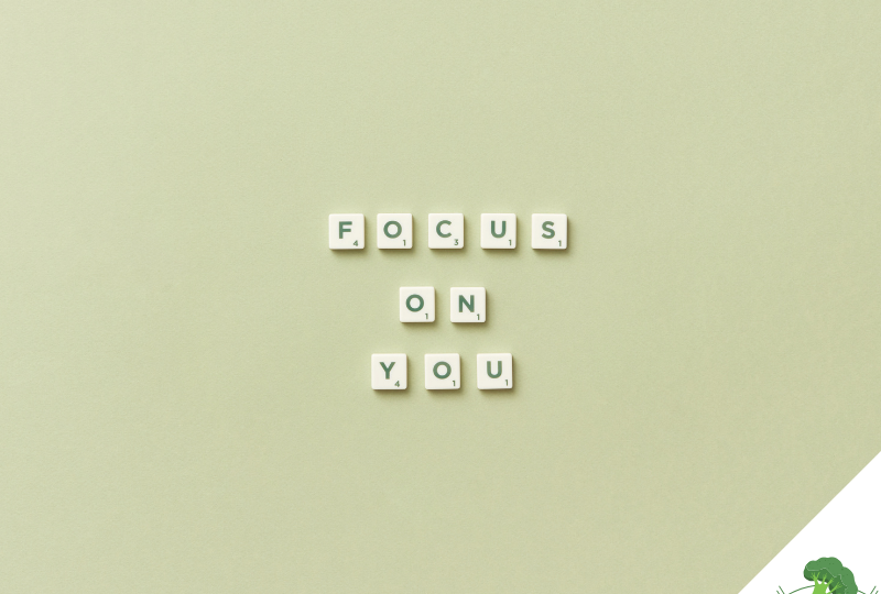 Scrabble-Buchstaben auf einem hellgrünen Hintergrund formen die Worte „FOCUS ON YOU“ in drei Zeilen, unten rechts befindet sich ein freier Bereich mit weißem Dreieck und einer Illustration eines grünen Brokkoli mit Hanteln.