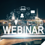 Eine Hand hält ein digitales Tablet, über dem das Wort „WEBINAR“ leuchtend erscheint. Darüber schweben zahlreiche Symbole wie Laptop, Person, E-Mail, Zahnräder und Glühbirne, die digitale Vernetzung und Online-Kommunikation symbolisieren. Im Hintergrund ist eine unscharfe Stadtansicht bei Nacht zu sehen.