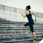 Frau in Sportkleidung läuft dynamisch eine breite Steintreppe hoch; sie trägt Sonnenbrille und Sportuhr, der Hintergrund zeigt eine moderne Betonfassade – Symbol für Aktivität, Fortschritt und körperliche Leistungsbereitschaft.