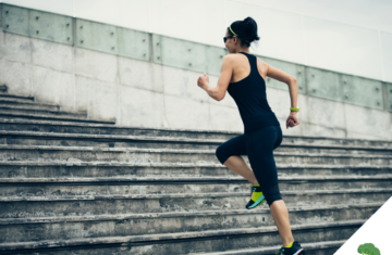 Frau in Sportkleidung läuft dynamisch eine breite Steintreppe hoch; sie trägt Sonnenbrille und Sportuhr, der Hintergrund zeigt eine moderne Betonfassade – Symbol für Aktivität, Fortschritt und körperliche Leistungsbereitschaft.