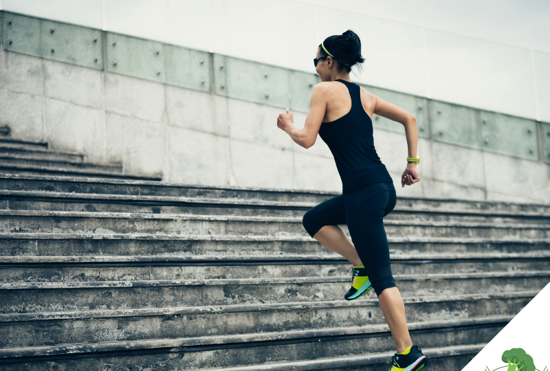 Frau in Sportkleidung läuft dynamisch eine breite Steintreppe hoch; sie trägt Sonnenbrille und Sportuhr, der Hintergrund zeigt eine moderne Betonfassade – Symbol für Aktivität, Fortschritt und körperliche Leistungsbereitschaft.