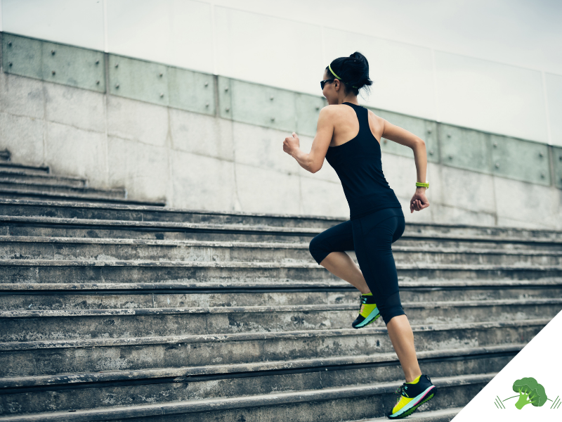 Frau in Sportkleidung läuft dynamisch eine breite Steintreppe hoch; sie trägt Sonnenbrille und Sportuhr, der Hintergrund zeigt eine moderne Betonfassade – Symbol für Aktivität, Fortschritt und körperliche Leistungsbereitschaft.