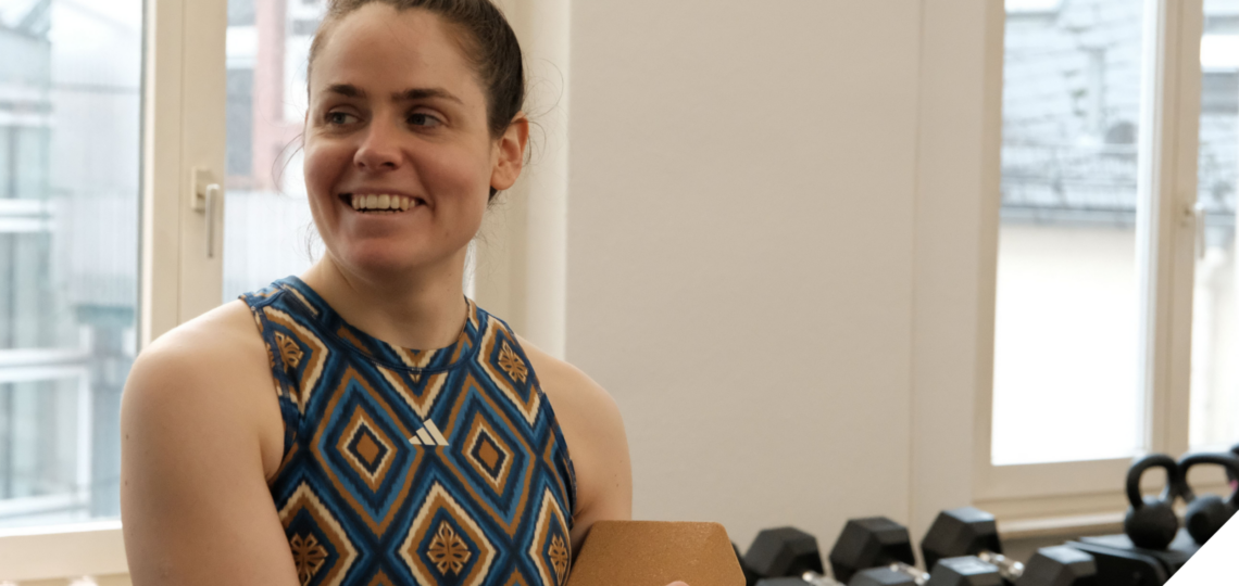 Eine Frau in sportlicher Kleidung steht in einem Fitnessraum vor einer Hantelablage, lächelt zur Seite und hält ein Klemmbrett in der Hand. Durch die Fenster im Hintergrund ist ein urbanes Gebäude zu sehen.