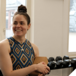 Eine Frau in sportlicher Kleidung steht in einem Fitnessraum vor einer Hantelablage, lächelt zur Seite und hält ein Klemmbrett in der Hand. Durch die Fenster im Hintergrund ist ein urbanes Gebäude zu sehen.