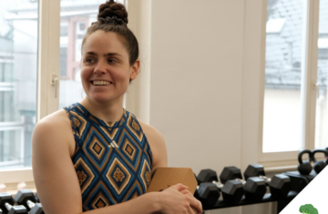 Eine Frau in sportlicher Kleidung steht in einem Fitnessraum vor einer Hantelablage, lächelt zur Seite und hält ein Klemmbrett in der Hand. Durch die Fenster im Hintergrund ist ein urbanes Gebäude zu sehen.