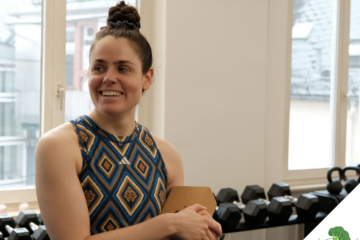 Eine Frau in sportlicher Kleidung steht in einem Fitnessraum vor einer Hantelablage, lächelt zur Seite und hält ein Klemmbrett in der Hand. Durch die Fenster im Hintergrund ist ein urbanes Gebäude zu sehen.