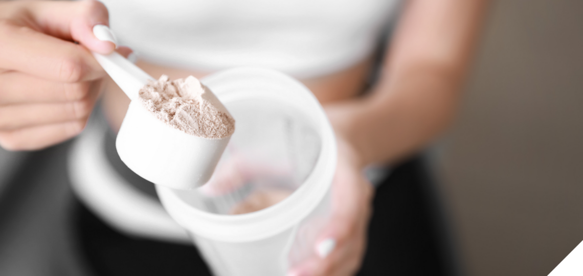 Nahaufnahme einer Frau, die Proteinpulver mit einem weißen Messlöffel in einen Shaker füllt. Sie trägt ein weißes Sporttop, der Hintergrund ist unscharf, wodurch der Fokus auf dem Pulver liegt.