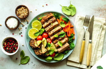 Ein bunt angerichteter veganer Teller mit mariniertem Tofu, frischen Limettenscheiben, Babyspinat, Karottenstreifen, Kirschtomaten und Sesam-Crackern, daneben kleine Schälchen mit Salz, Pfeffer und Koriandersamen sowie Besteck auf einer Leinenserviette.