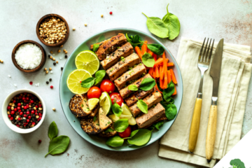 Ein bunt angerichteter veganer Teller mit mariniertem Tofu, frischen Limettenscheiben, Babyspinat, Karottenstreifen, Kirschtomaten und Sesam-Crackern, daneben kleine Schälchen mit Salz, Pfeffer und Koriandersamen sowie Besteck auf einer Leinenserviette.