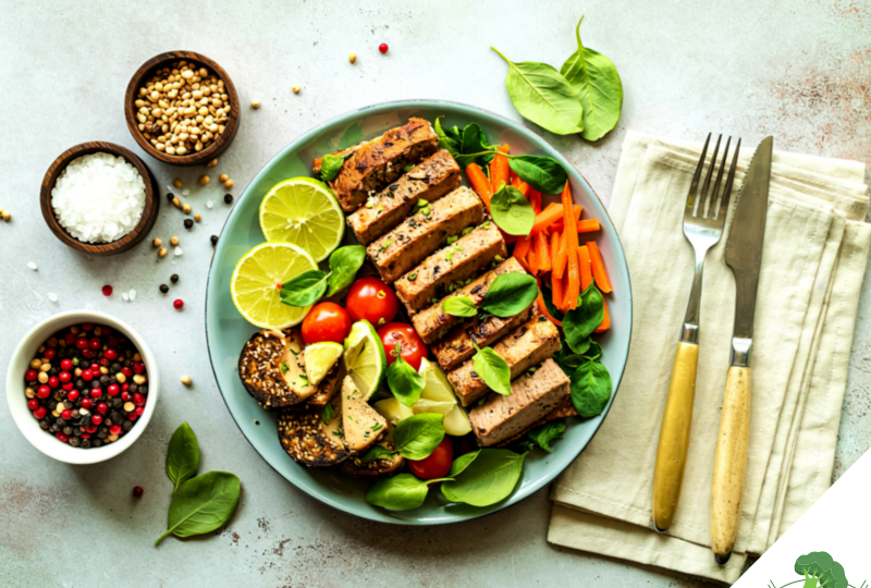 Ein bunt angerichteter veganer Teller mit mariniertem Tofu, frischen Limettenscheiben, Babyspinat, Karottenstreifen, Kirschtomaten und Sesam-Crackern, daneben kleine Schälchen mit Salz, Pfeffer und Koriandersamen sowie Besteck auf einer Leinenserviette.