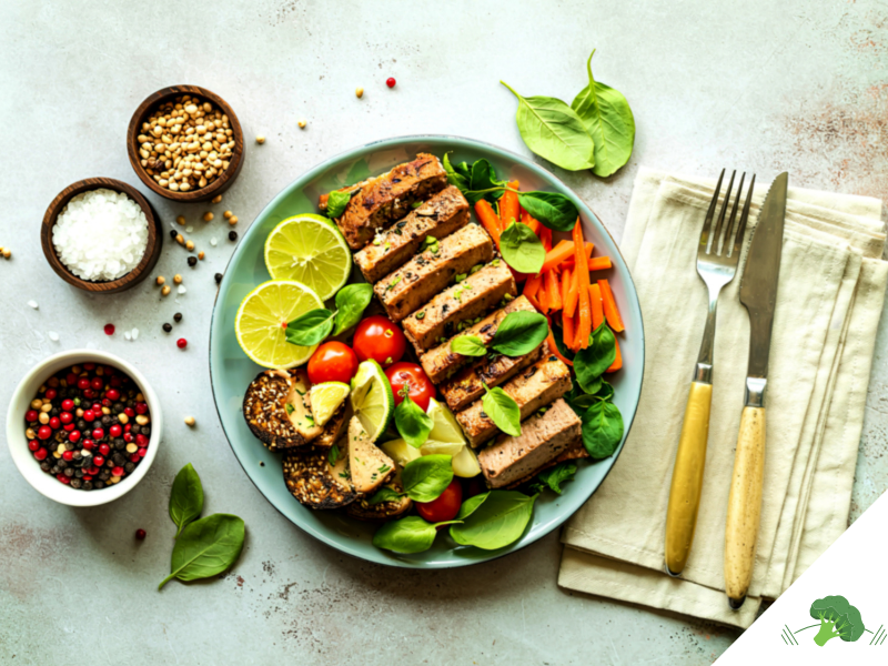 Ein bunt angerichteter veganer Teller mit mariniertem Tofu, frischen Limettenscheiben, Babyspinat, Karottenstreifen, Kirschtomaten und Sesam-Crackern, daneben kleine Schälchen mit Salz, Pfeffer und Koriandersamen sowie Besteck auf einer Leinenserviette.