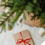 Ein festlich verpacktes Geschenk mit rotem Band liegt auf einem weißen, flauschigen Teppich unter einem geschmückten Weihnachtsbaum mit goldenen Kugeln und warmem Licht.