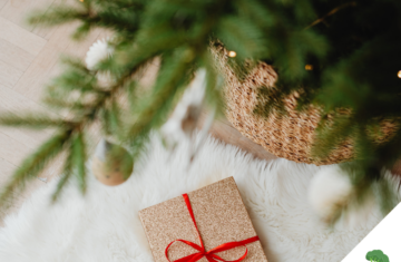 Ein festlich verpacktes Geschenk mit rotem Band liegt auf einem weißen, flauschigen Teppich unter einem geschmückten Weihnachtsbaum mit goldenen Kugeln und warmem Licht.