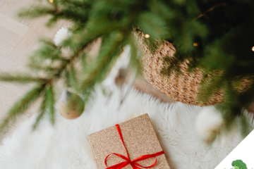 Ein festlich verpacktes Geschenk mit rotem Band liegt auf einem weißen, flauschigen Teppich unter einem geschmückten Weihnachtsbaum mit goldenen Kugeln und warmem Licht.