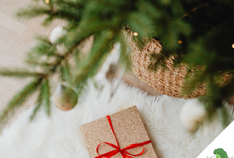 Ein festlich verpacktes Geschenk mit rotem Band liegt auf einem weißen, flauschigen Teppich unter einem geschmückten Weihnachtsbaum mit goldenen Kugeln und warmem Licht.