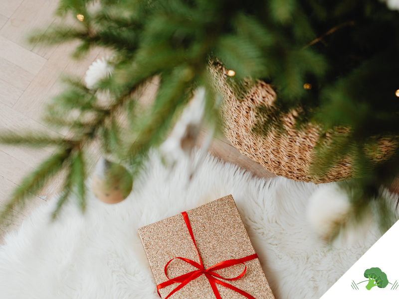 Ein festlich verpacktes Geschenk mit rotem Band liegt auf einem weißen, flauschigen Teppich unter einem geschmückten Weihnachtsbaum mit goldenen Kugeln und warmem Licht.