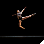 Eine Tänzerin in sportlicher Kleidung springt anmutig in der Luft auf einer dunklen Bühne, mit ausgestreckten Armen und Beinen – voller Kraft und Leichtigkeit.