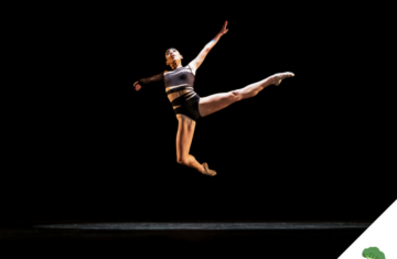 Eine Tänzerin in sportlicher Kleidung springt anmutig in der Luft auf einer dunklen Bühne, mit ausgestreckten Armen und Beinen – voller Kraft und Leichtigkeit.