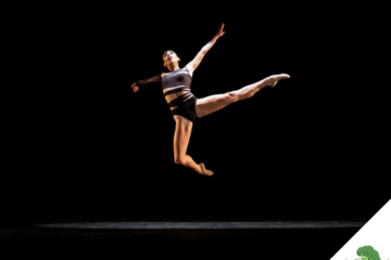 Eine Tänzerin in sportlicher Kleidung springt anmutig in der Luft auf einer dunklen Bühne, mit ausgestreckten Armen und Beinen – voller Kraft und Leichtigkeit.