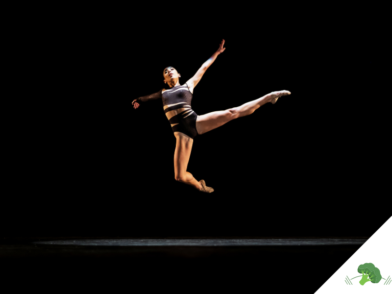 Eine Tänzerin in sportlicher Kleidung springt anmutig in der Luft auf einer dunklen Bühne, mit ausgestreckten Armen und Beinen – voller Kraft und Leichtigkeit.