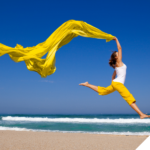 Frau springt kraftvoll und frei am Strand in die Luft, ein leuchtend gelbes Tuch weht dynamisch über ihr – Symbol für Leichtigkeit, Energie und die befreiende Wirkung eines beschwerdefreien Körpers.