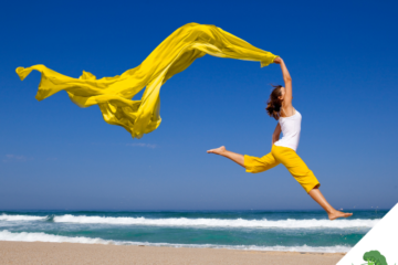 Frau springt kraftvoll und frei am Strand in die Luft, ein leuchtend gelbes Tuch weht dynamisch über ihr – Symbol für Leichtigkeit, Energie und die befreiende Wirkung eines beschwerdefreien Körpers.
