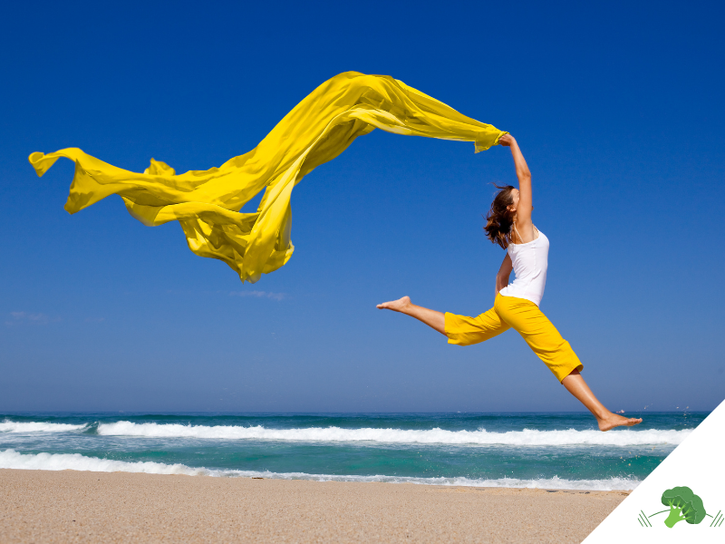 Frau springt kraftvoll und frei am Strand in die Luft, ein leuchtend gelbes Tuch weht dynamisch über ihr – Symbol für Leichtigkeit, Energie und die befreiende Wirkung eines beschwerdefreien Körpers.