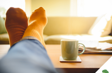 Person mit orangefarbenen Socken und blauer Jeans hat die Füße entspannt auf einem Holztisch hochgelegt, daneben steht eine dampfende Tasse auf einem Untersetzer, im Hintergrund ein gemütliches Sofa im Sonnenlicht.