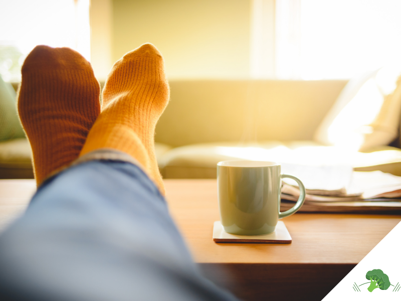 Person mit orangefarbenen Socken und blauer Jeans hat die Füße entspannt auf einem Holztisch hochgelegt, daneben steht eine dampfende Tasse auf einem Untersetzer, im Hintergrund ein gemütliches Sofa im Sonnenlicht.