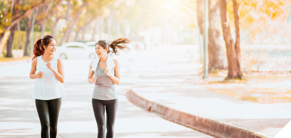 Zwei sportlich gekleidete Frauen joggen lachend nebeneinander auf einem von Bäumen gesäumten Weg in herbstlicher Atmosphäre. Die Sonne scheint durch das Laub, wodurch eine warme, einladende Stimmung entsteht.