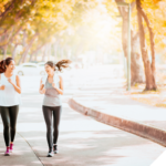 Zwei sportlich gekleidete Frauen joggen lachend nebeneinander auf einem von Bäumen gesäumten Weg in herbstlicher Atmosphäre. Die Sonne scheint durch das Laub, wodurch eine warme, einladende Stimmung entsteht.